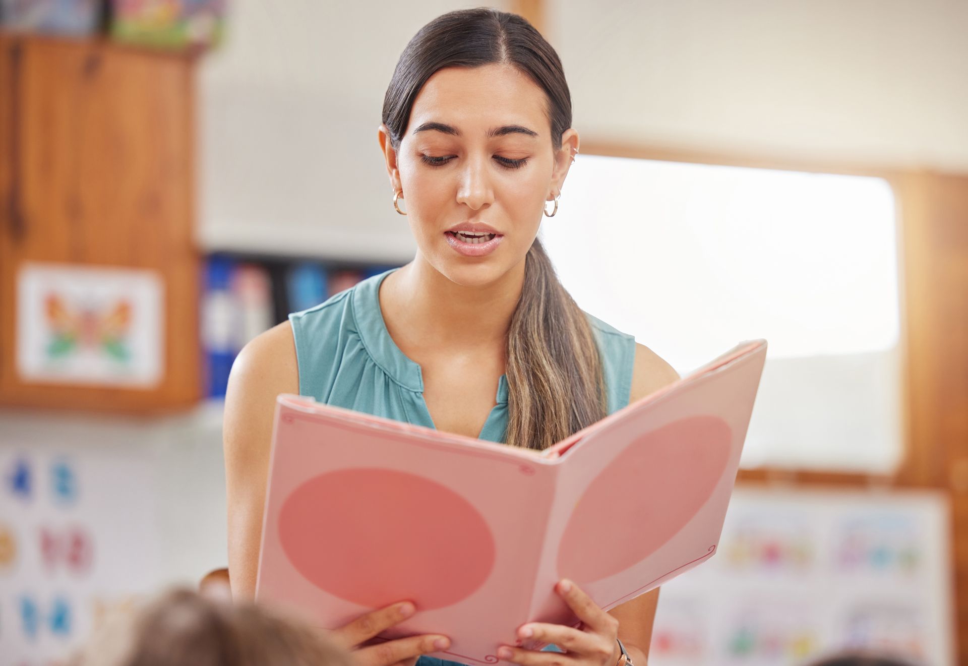 Woman reading a pink book aloud in a classroom, wearing a teal top.