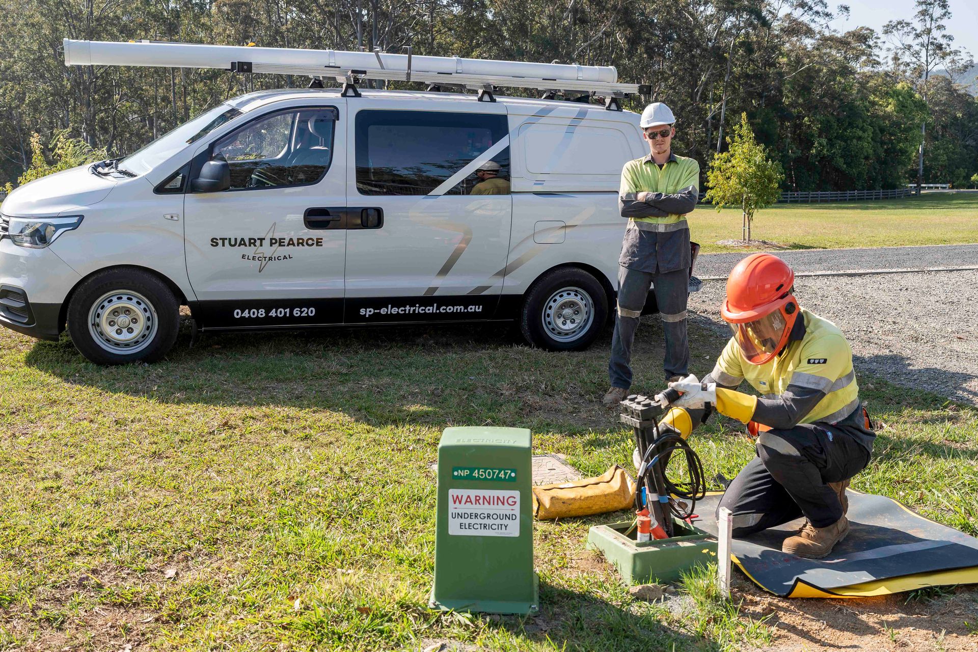 Two utility workers in safety gear near a van, one kneeling at a green box, the other standing — Stuart Pearce Electrical In NSW Coffs Harbour, NSW