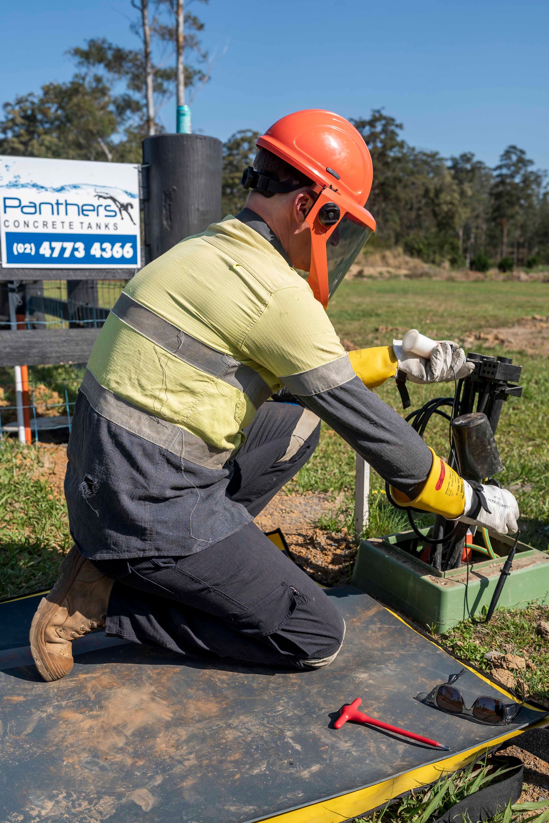 Worker kneels, adjusting equipment outdoors, wearing a hard hat, face shield, and protective gear — Stuart Pearce Electrical In NSW Sawtell, NSW