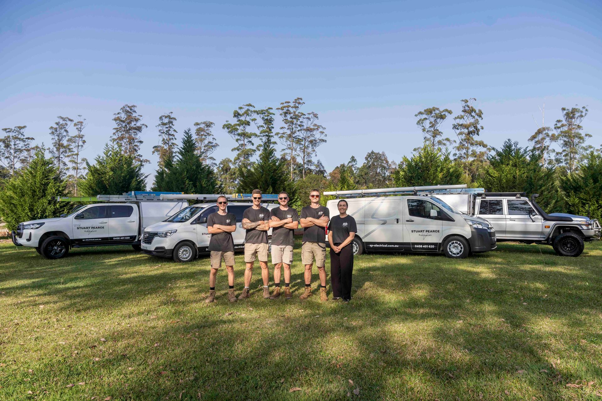 Team of six people in front of five work vehicles on a grassy lawn — Stuart Pearce Electrical In Coffs Harbour, NSW