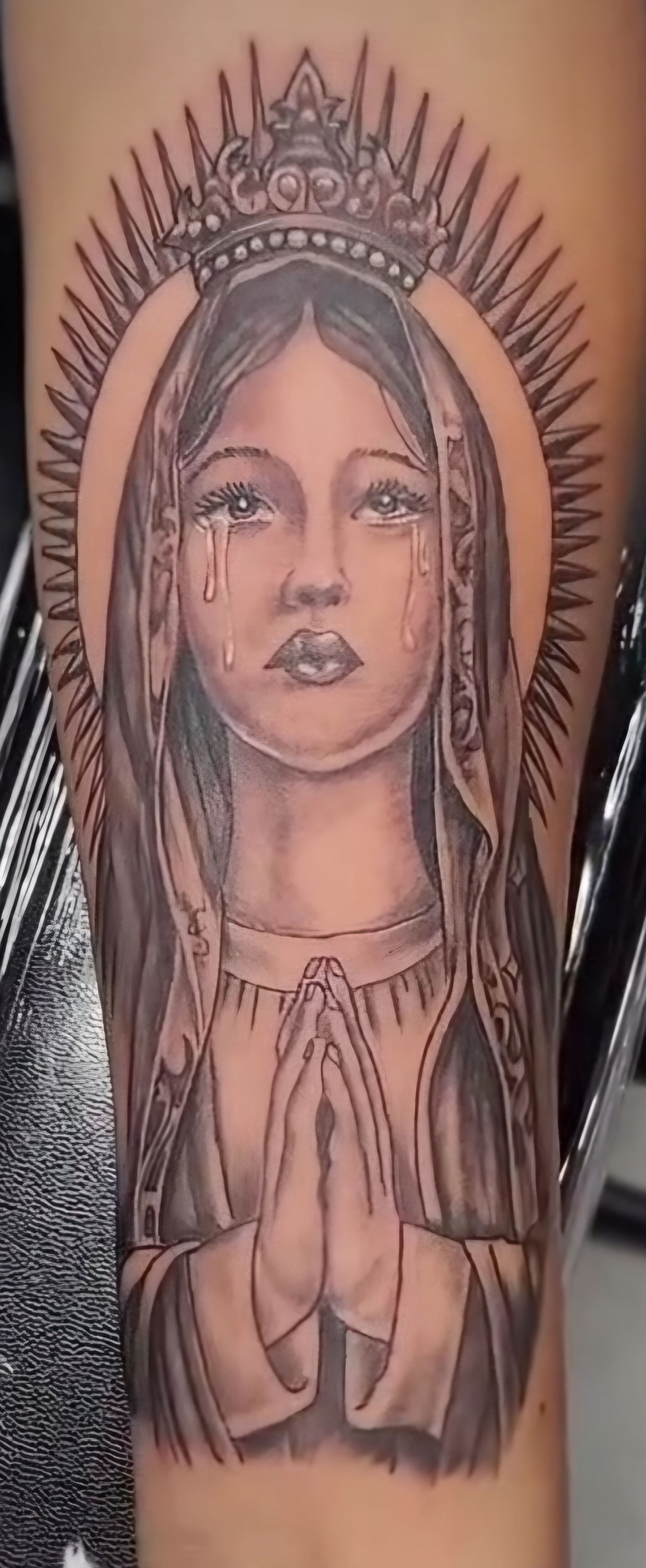 Black and gray tattoo of the Virgin Mary with a crown, halo, tears, and praying hands.