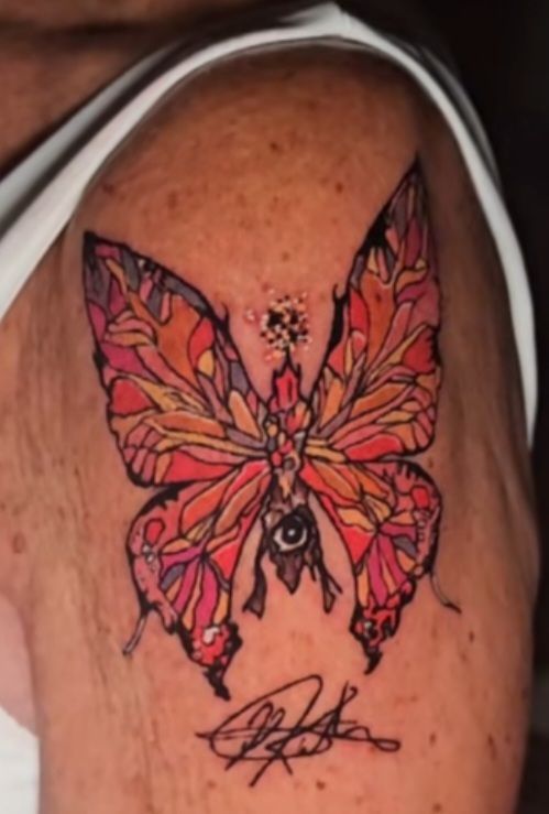 Colorful butterfly tattoo on a person's shoulder, with an eye at the body and signature below.