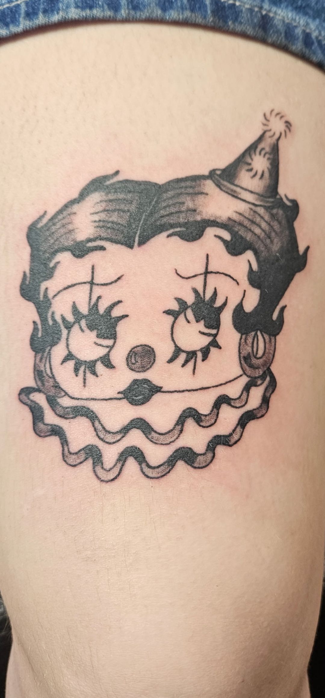 Black-and-white clown face tattoo on skin, wearing a pointed party hat with ruffled collar.
