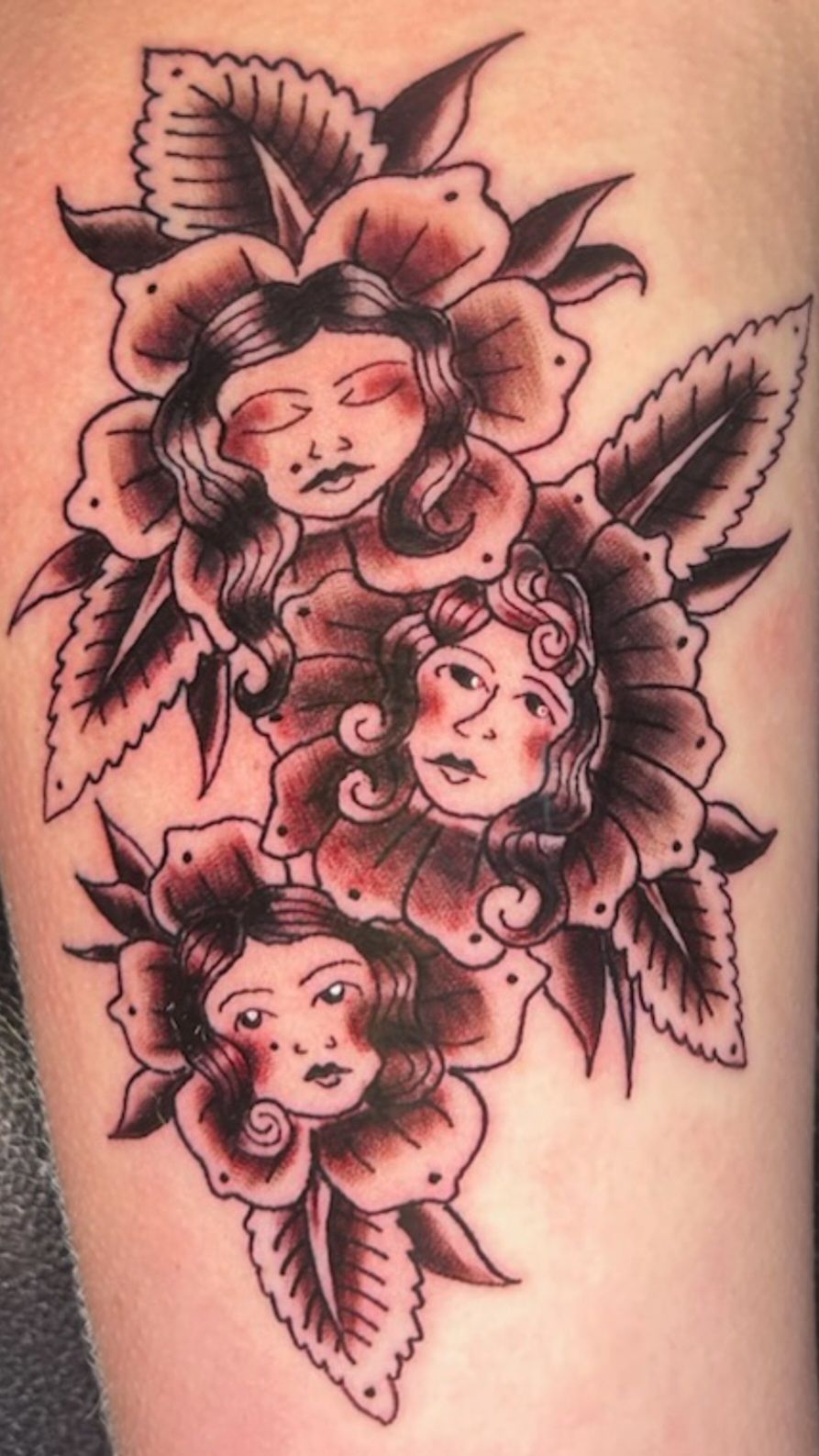 Three black-and-gray tattooed cherub faces surrounded by leaves on skin.
