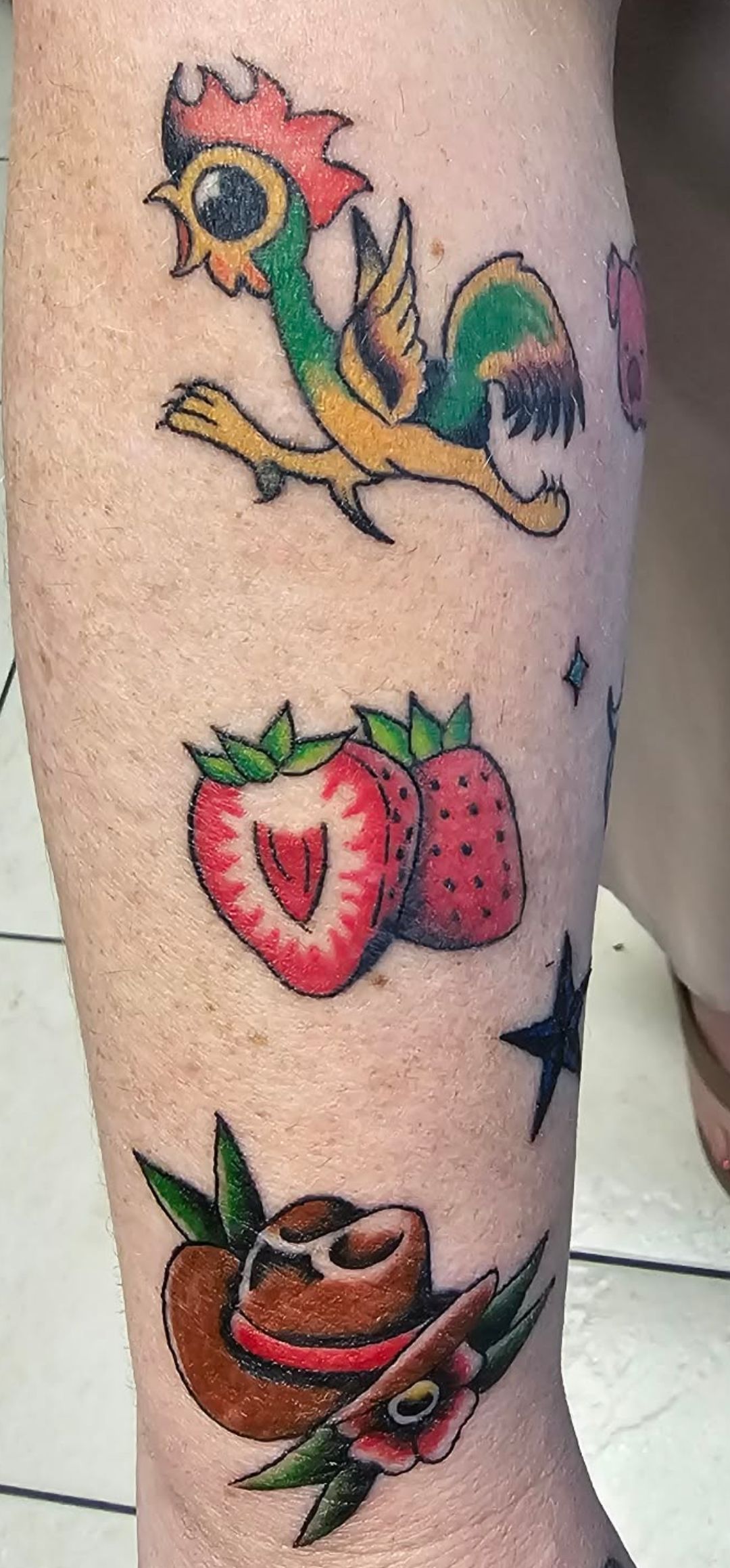 Three colorful tattoo designs on a leg: a rooster, strawberries, and a bowl with fruit and flowers.