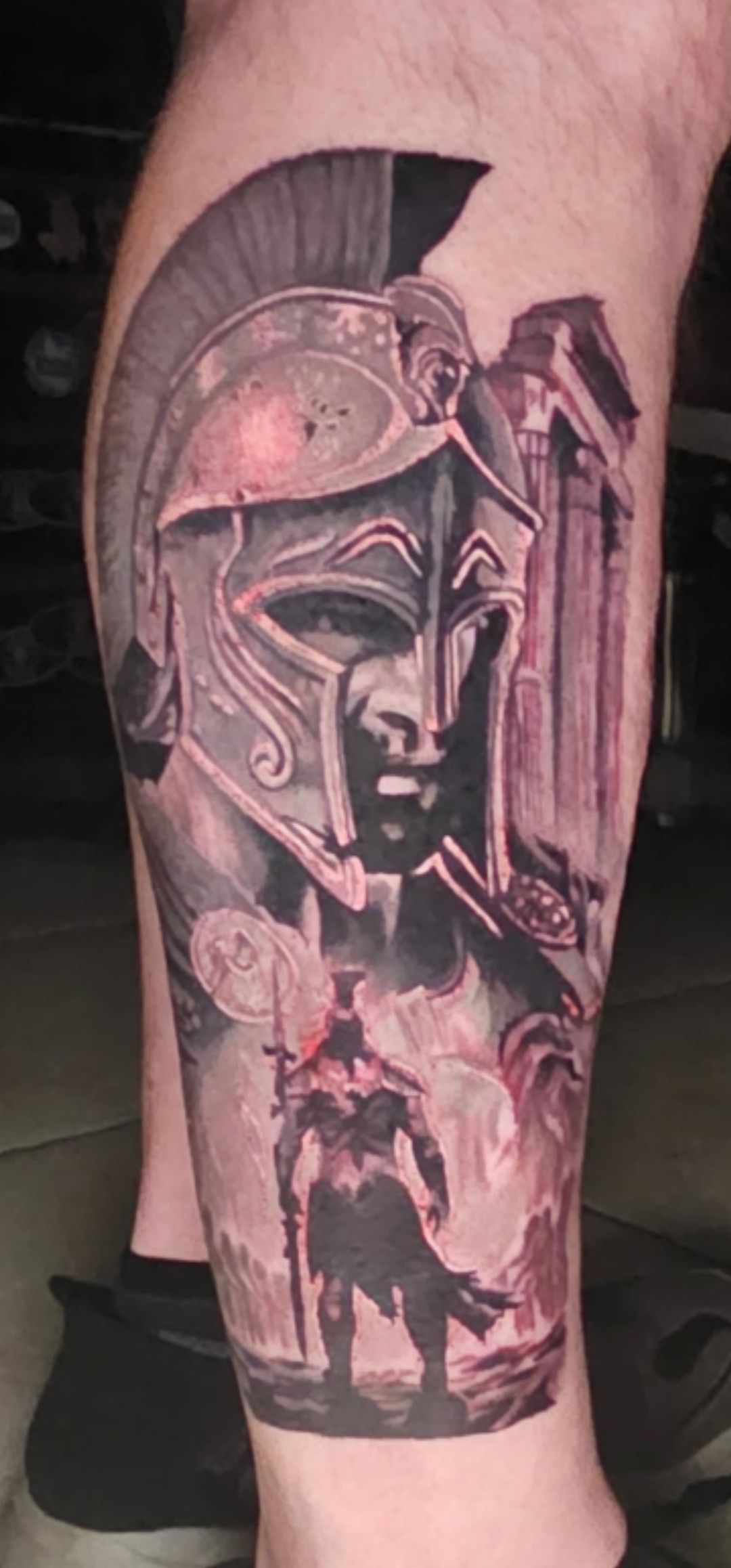 Black-and-pink tattoo of a Spartan helmet and soldier on a calf, with a church in the background