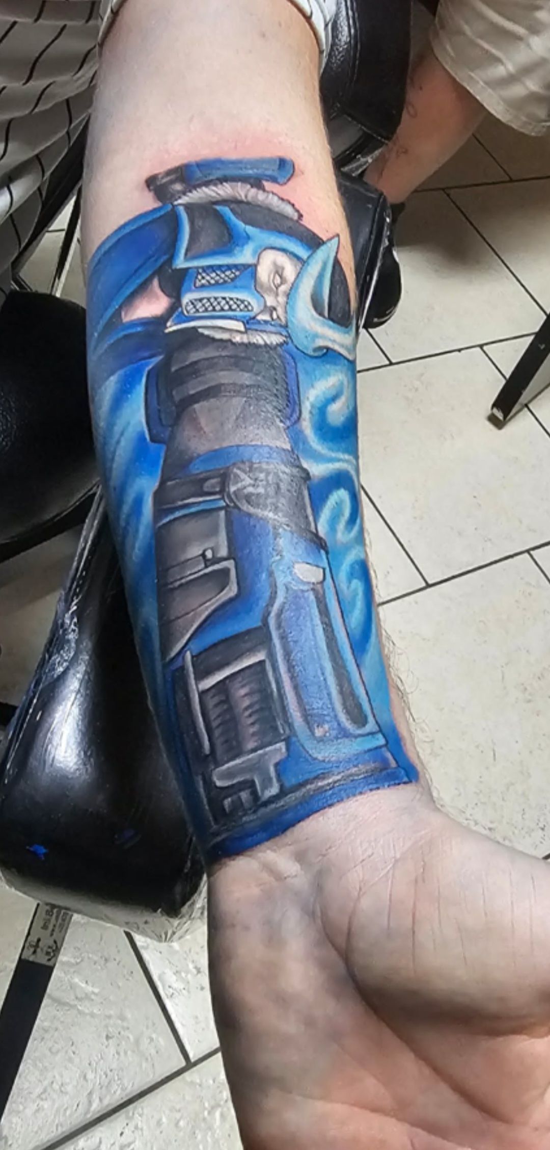 Blue mechanical-style tattoo covering a forearm, shown with the wrist turned upward on a chair.