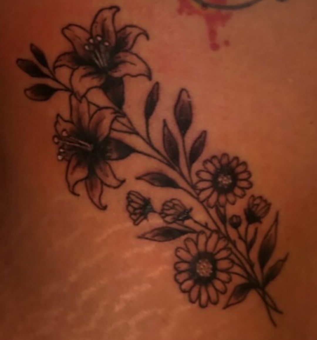 Black floral tattoo with lilies, daisies, leaves, and a butterfly on skin