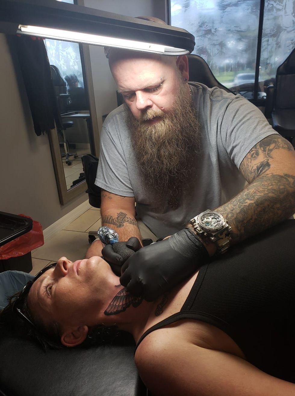 A man with a beard is tattooing a woman 's neck.