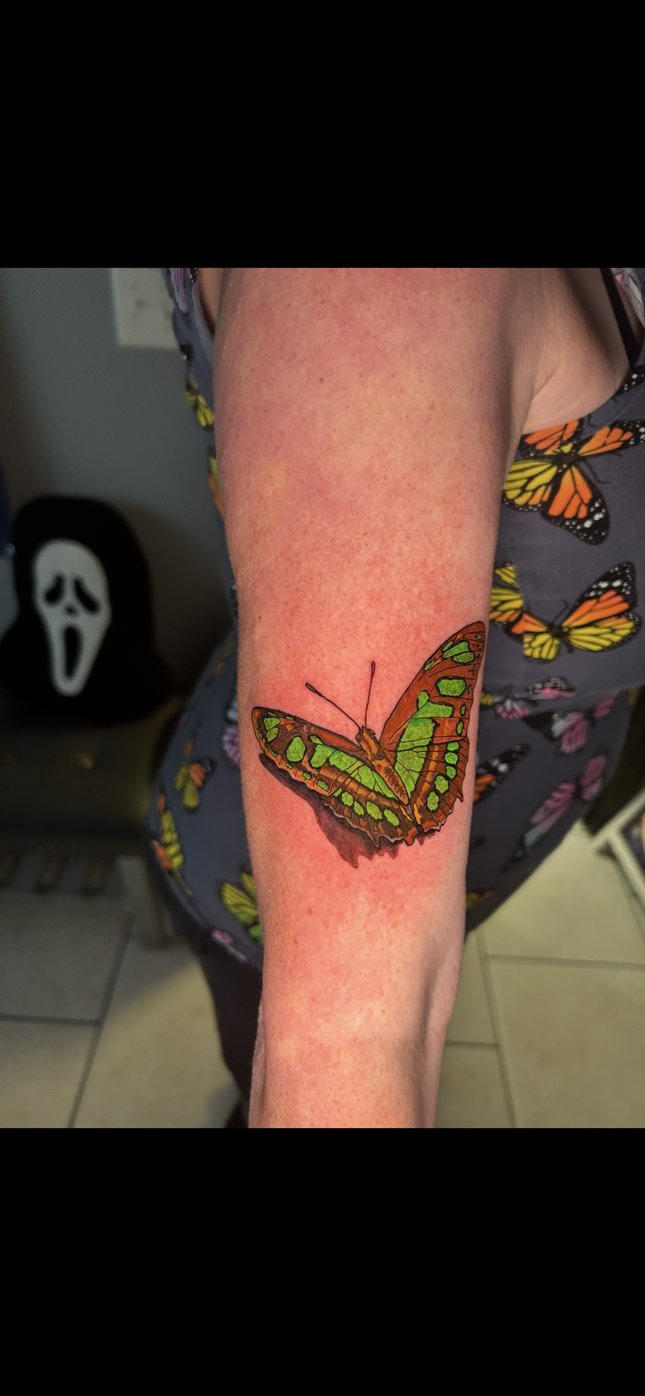 A person's arm with a green and brown butterfly tattoo. A Scream mask is in the background.