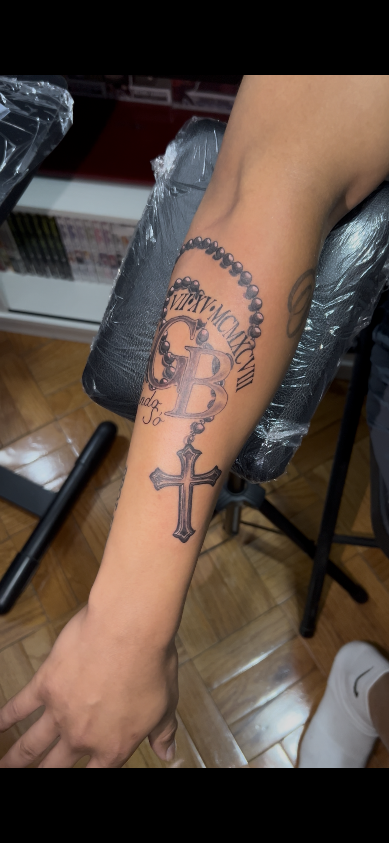 Arm with a rosary and cross tattoo. The tattoo is black and gray. The arm is resting on a black surface.