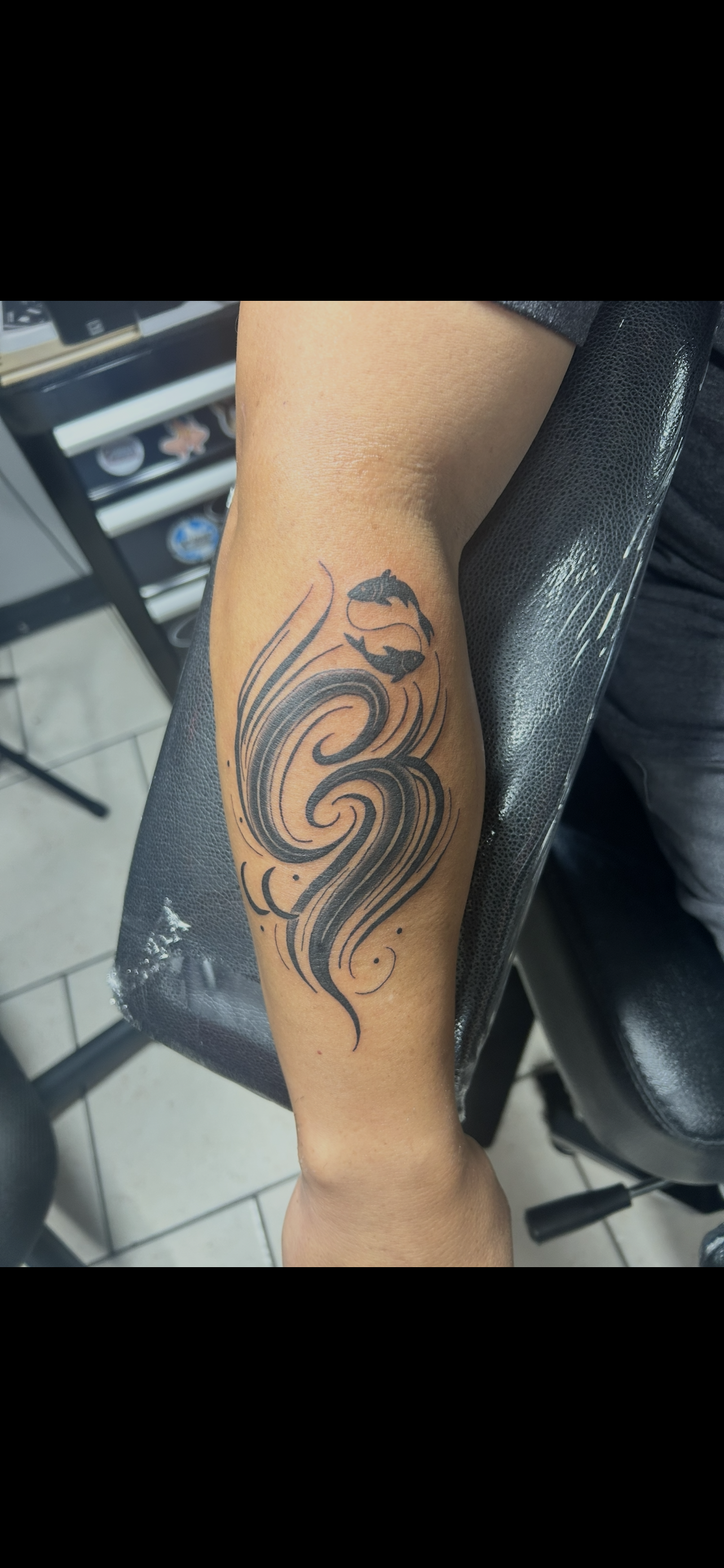 A tattoo on a forearm, with black swirling tribal designs. The arm rests on a black chair.