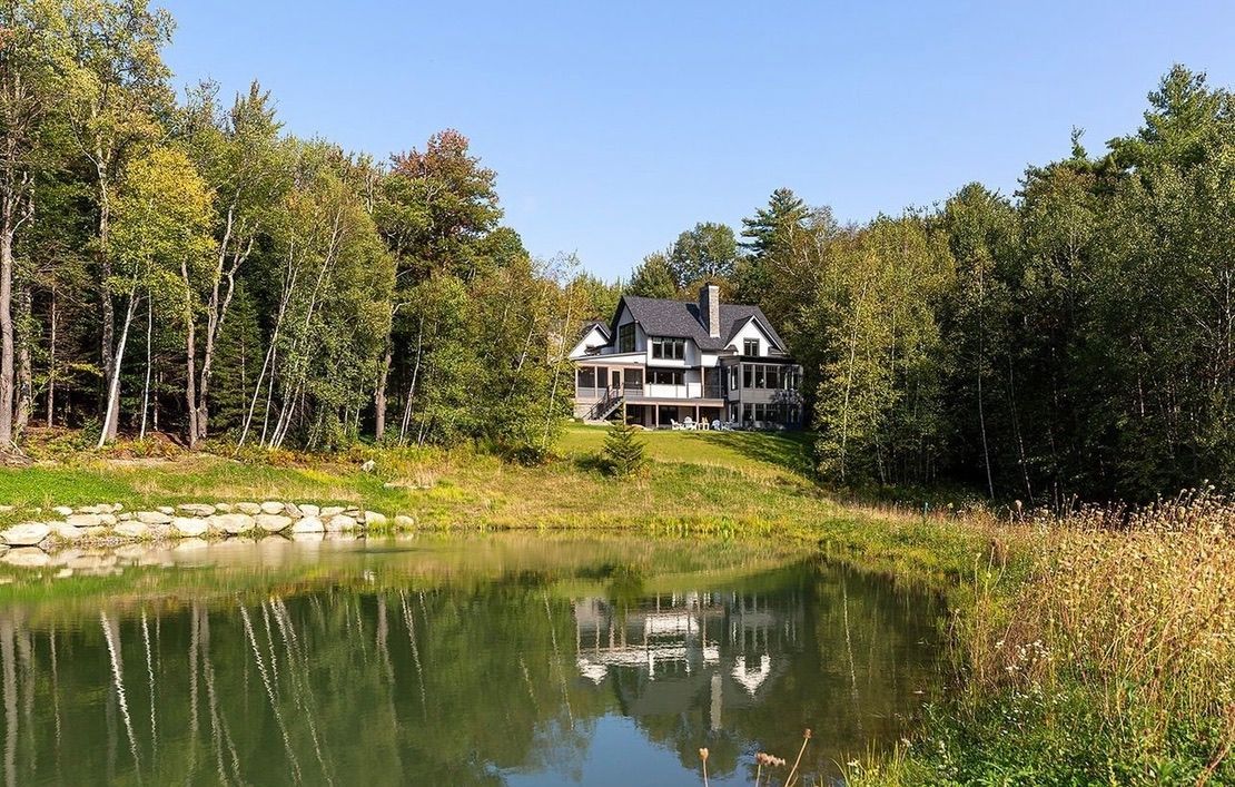 Lakeside house reflected in a calm pond, surrounded by green trees under a clear blue sky