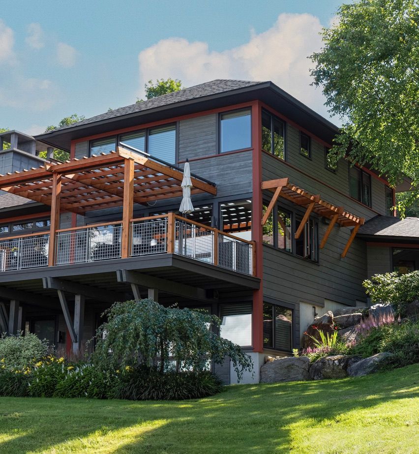 Modern two-story house with dark wood siding, large deck, and green lawn on a sunny hillside