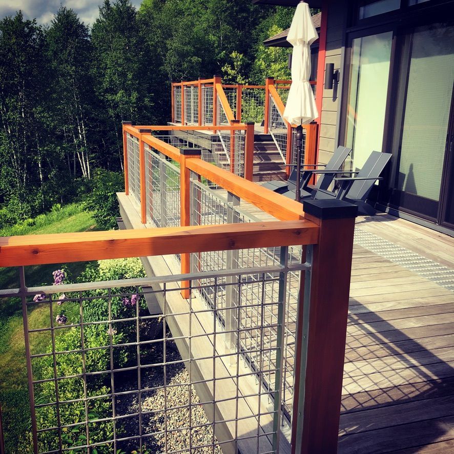 Wooden deck with glass railings overlooking lush greenery, with patio chairs and a closed umbrella.