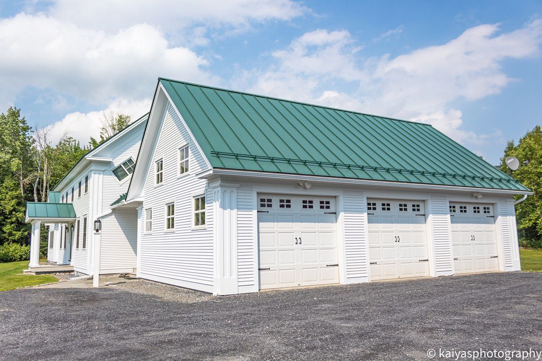 White house with green metal roof and three garage doors on a gravel driveway under a blue sky