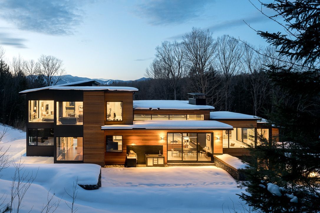 Snowy modern house at dusk with warm lights glowing through large windows.