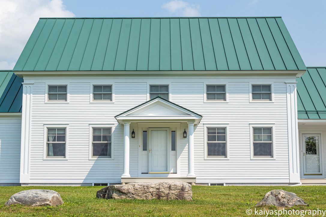 White farmhouse with a green metal roof and small front porch, set on a grassy lawn.