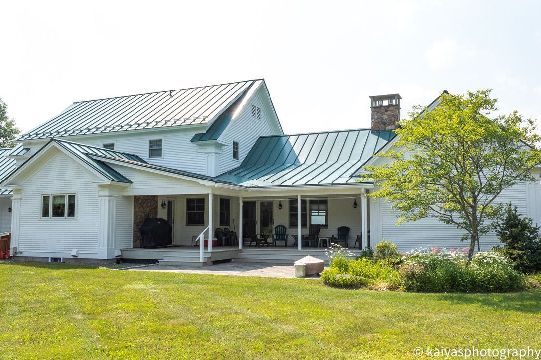 White house with a metal roof and covered porch, set on a grassy lawn with trees and shrubs.