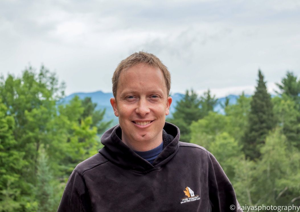 Duane smiling in a dark hoodie outdoors with green trees and mountains in the background