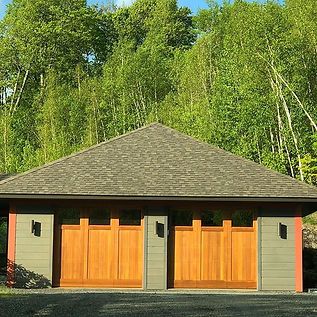 Single-story garage with wooden double doors and a dark roof, set against green trees.