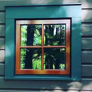 Wooden-framed window on teal siding reflecting green trees and a forest scene