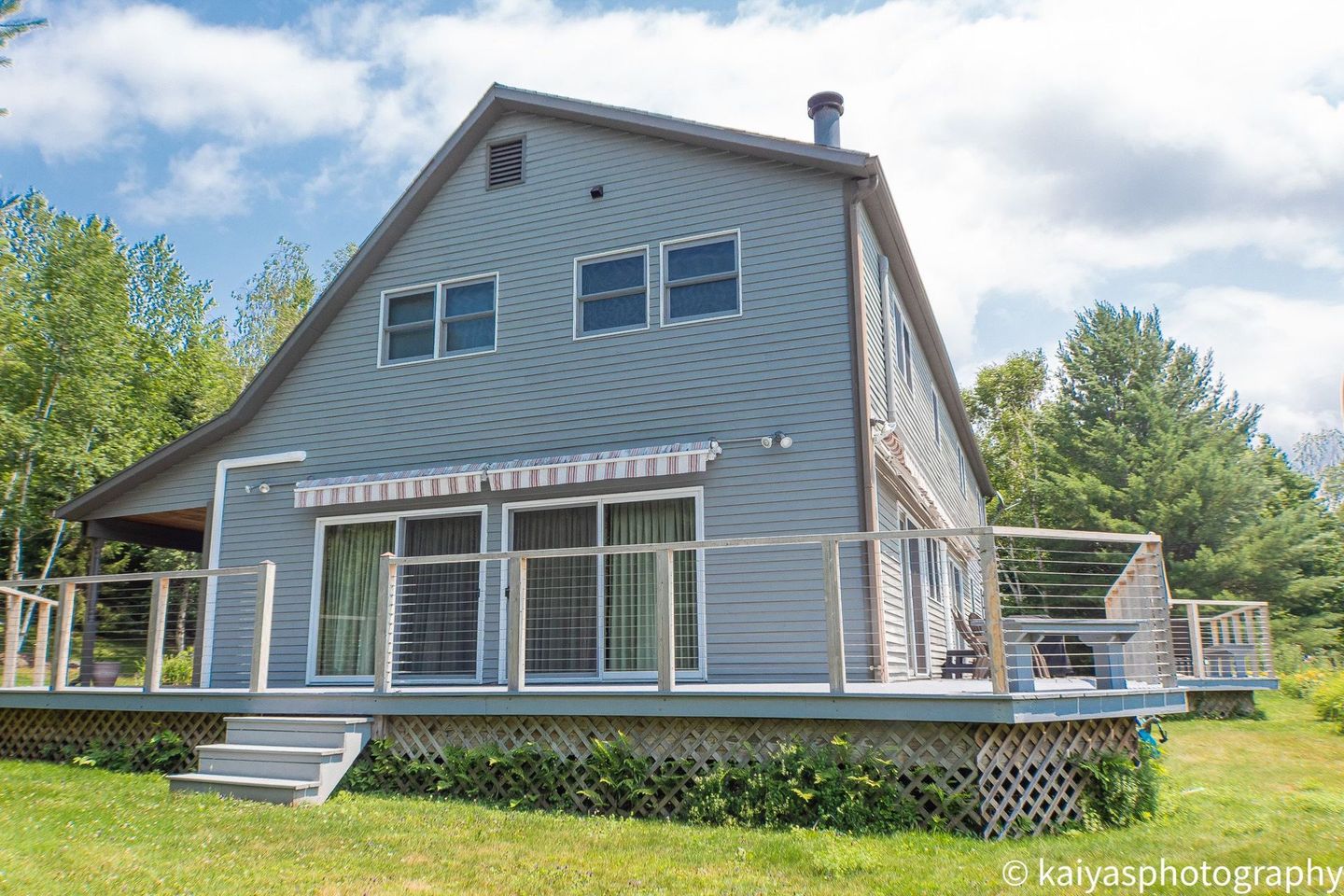 Gray two-story house with a wraparound deck and large windows, surrounded by trees and green lawn.