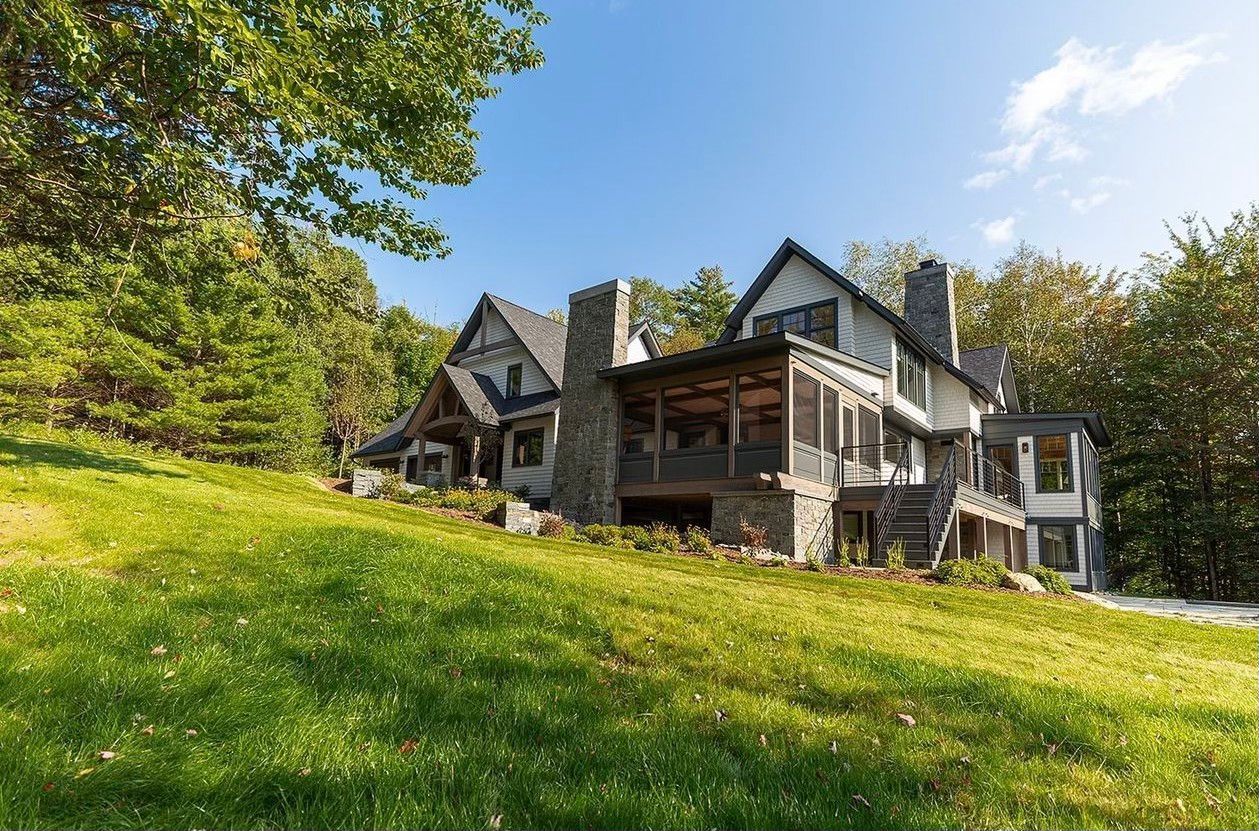 Large rustic house on a grassy hillside under a blue sky, surrounded by trees