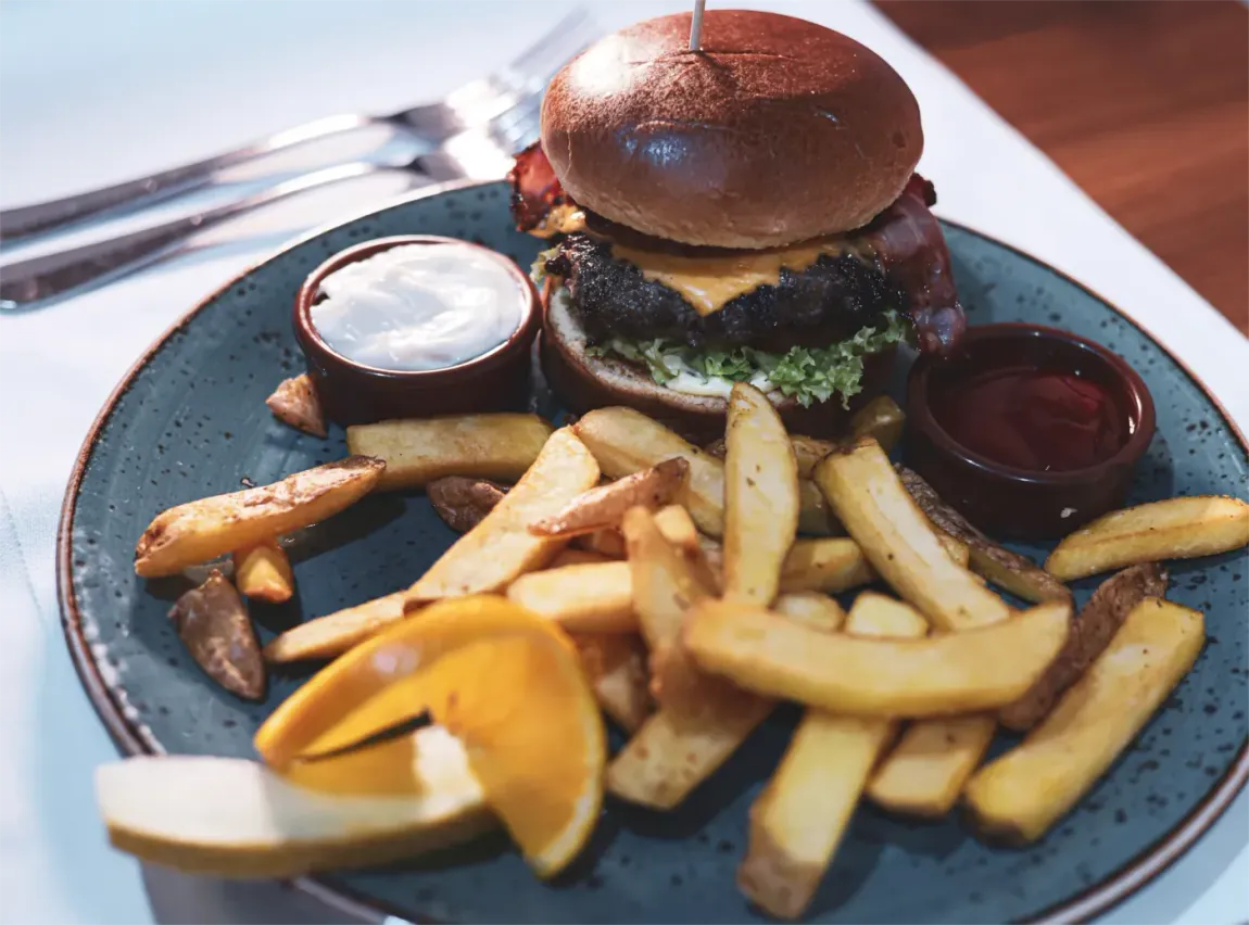 Burger mit Pommes frites, zwei Dips und einer Garnitur auf einem blauen Teller, im Hintergrund Besteck.
