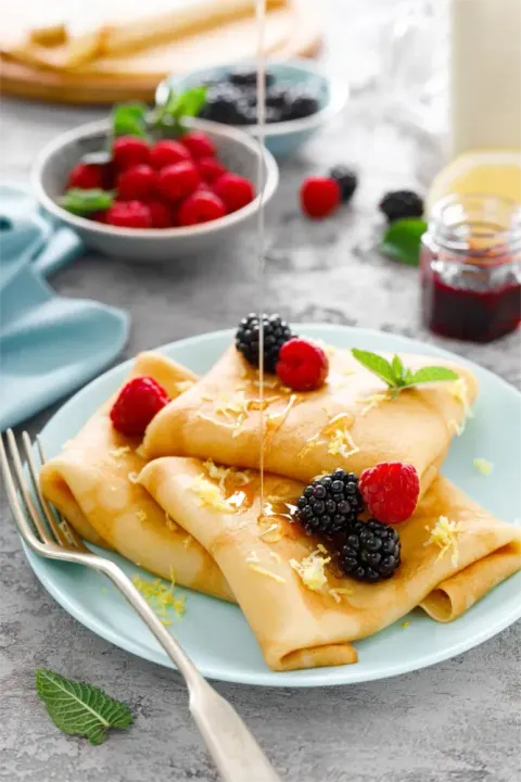 Crêpes auf einem blauen Teller, garniert mit Beeren, Zitronenschale und beträufeltem Honig.