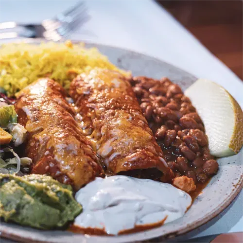 Teller mit Enchiladas, gelbem Reis, Bohnen, Guacamole, Sauce und einer Melonenscheibe.