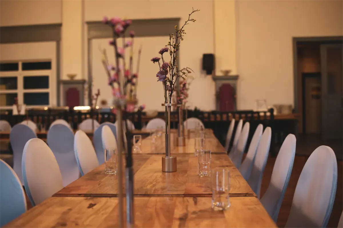 Lange Holztische mit Gläsern und Blumengestecken, weiß bezogene Stühle in Reihen in einer großen Halle.