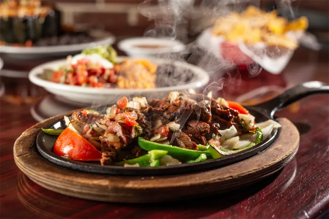 Zischende Fajitas mit Gemüse in einer gusseisernen Pfanne. Im Hintergrund auf einem Holztisch weitere mexikanische Gerichte.