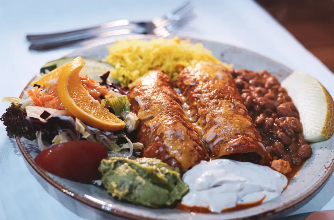 Teller mit Enchiladas, gelbem Reis, Bohnen, Salat, Guacamole, Orangenscheibe und Sauerrahm.