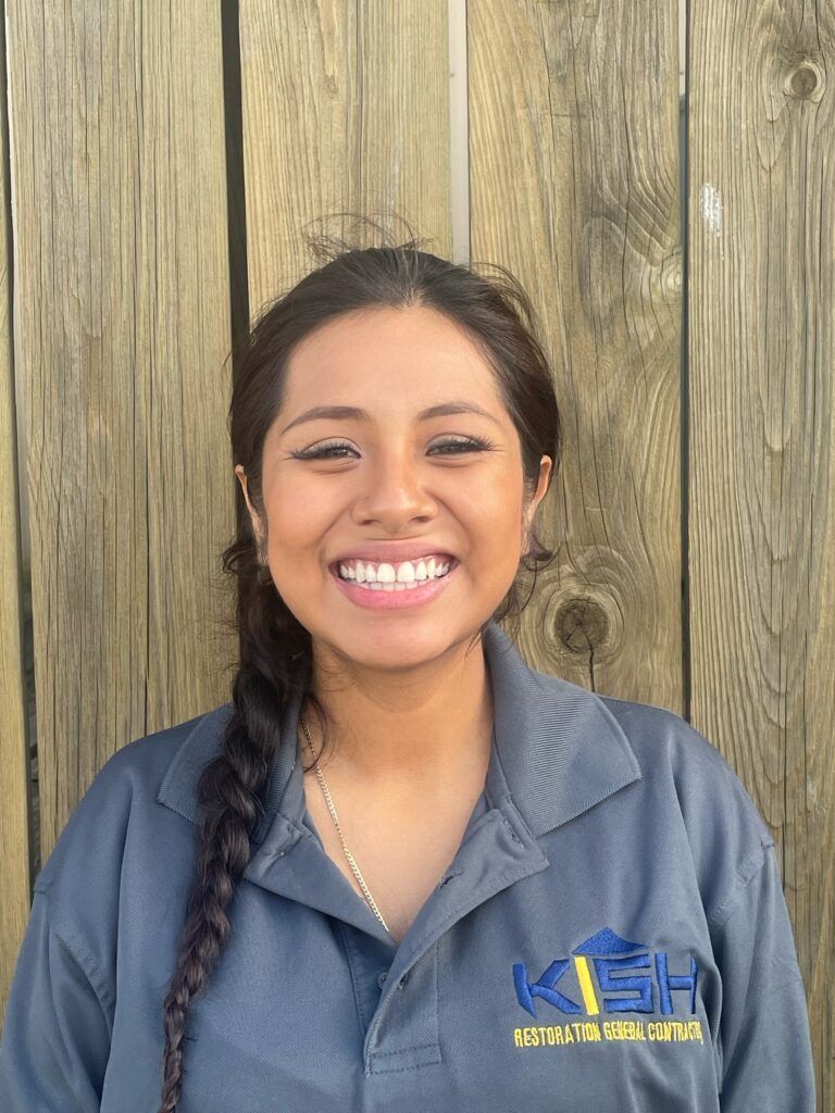 A woman in a blue shirt is smiling in front of a wooden wall.