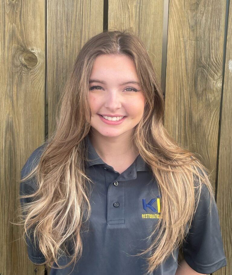 A young woman wearing a gray polo shirt is smiling in front of a wooden fence.