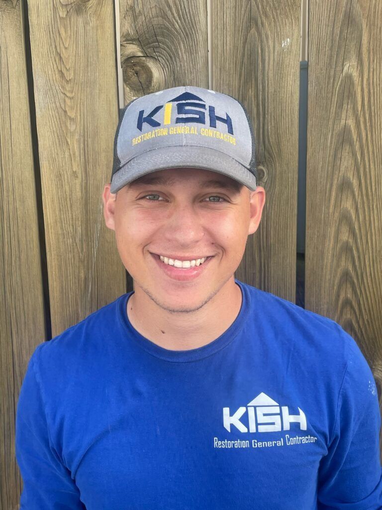 A man wearing a hat and a blue shirt is smiling in front of a wooden fence.