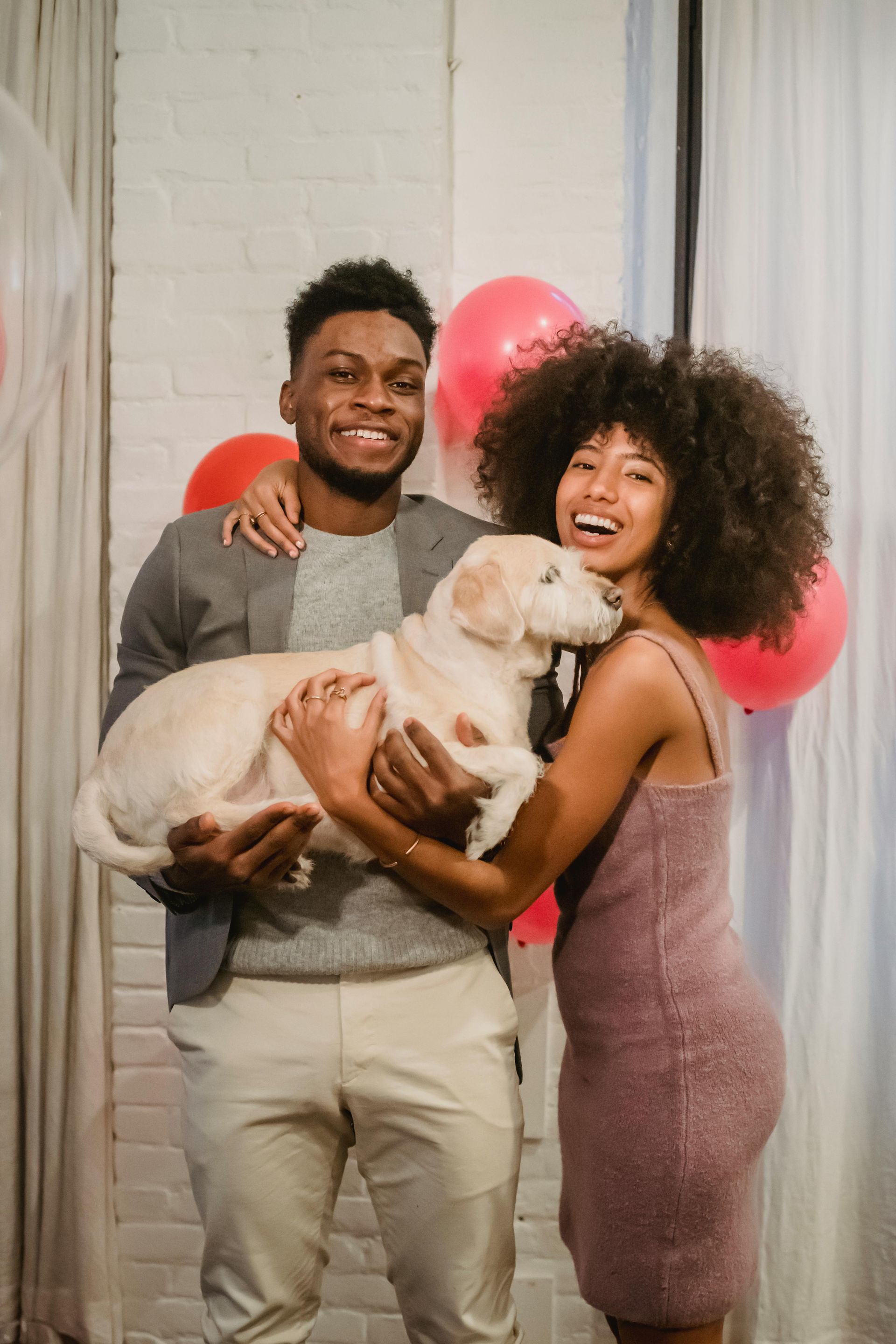 Couple holding a light-colored dog, smiling with balloons in the background.