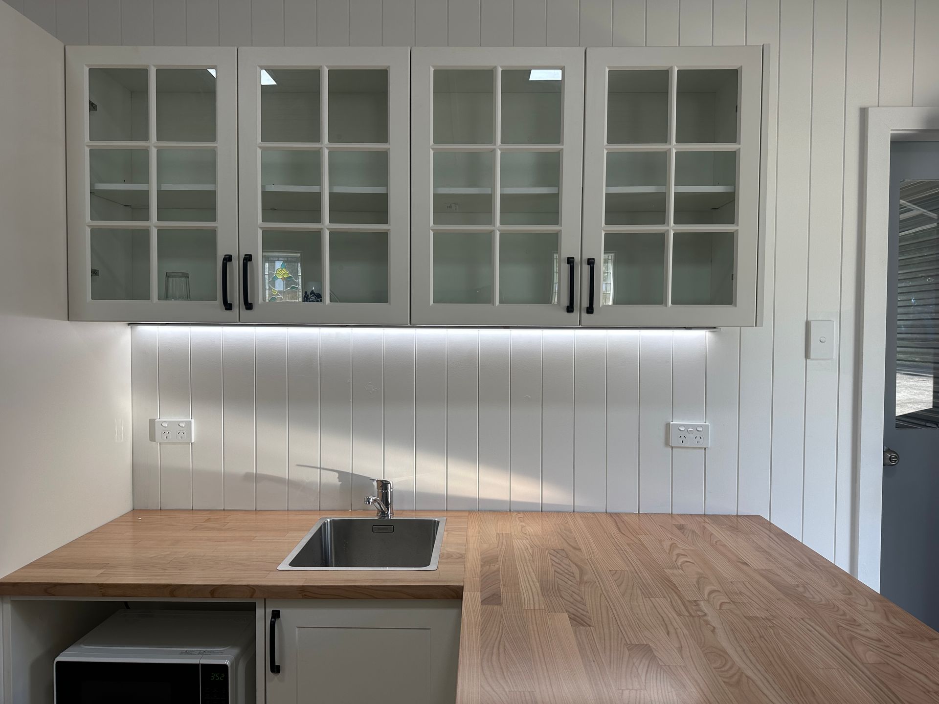 White Kitchen Cabinets With Glass Doors Over a Wood Countertop, Sink, and Backsplash — SparkSafe Electrical in Dapto, NSW