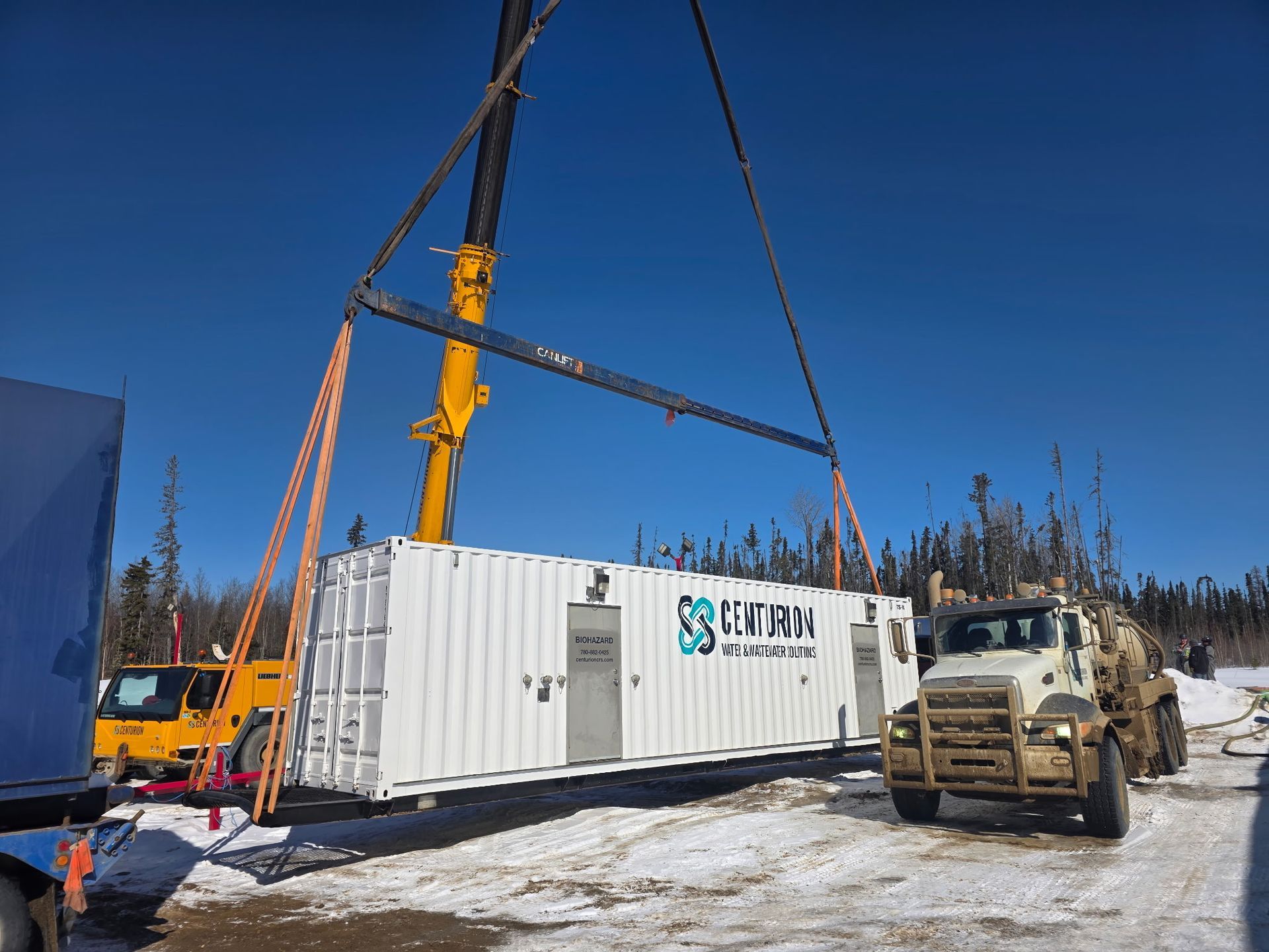 Crane lifting a white shipping container at a construction site, with an excavator nearby.
