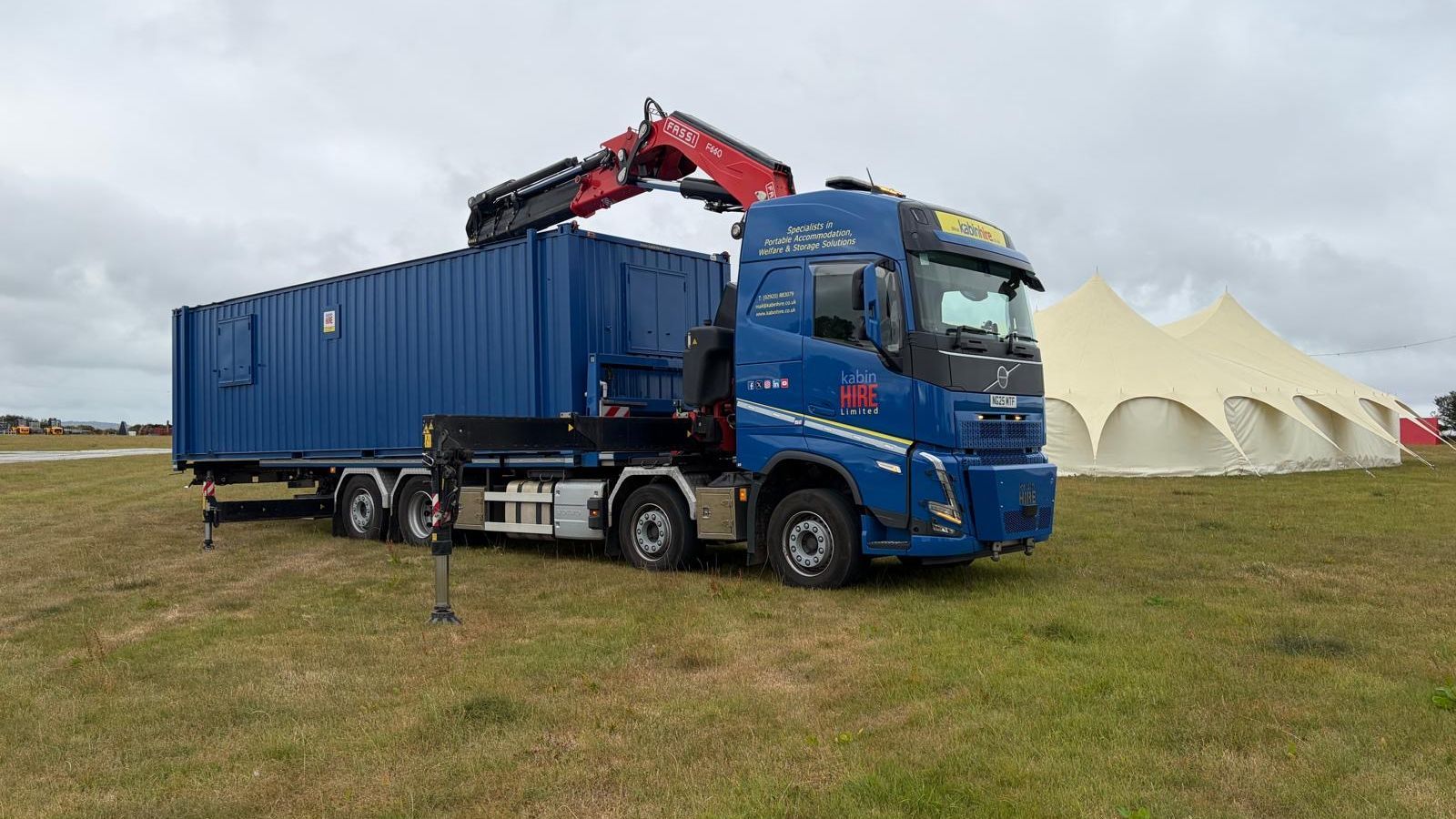 Blue Centurion Kabin Hire truck and red crane hauling a blue container in a field near white tent.
