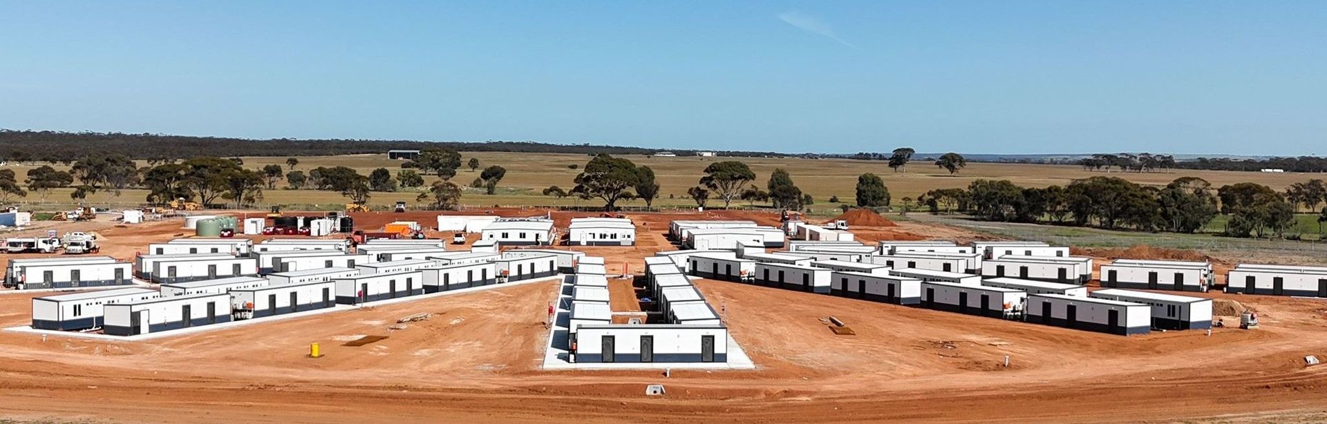 Aerial view of prefabricated buildings arranged on a brown field under a clear blue sky.