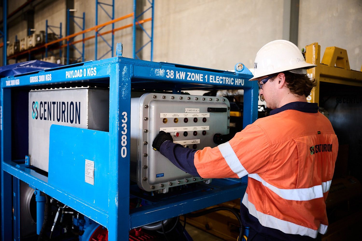 A worker in an orange shirt and white hard hat operates electrical equipment on a blue metal frame.
