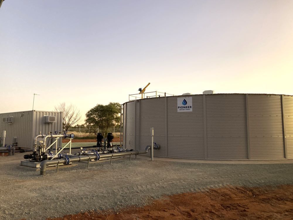Water storage tank and industrial building in an outdoor setting with gravel ground under a clear sky.