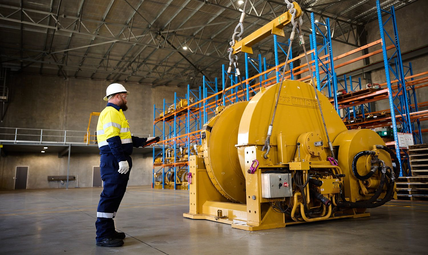Worker in safety gear operates crane to lift large yellow machinery in a warehouse.