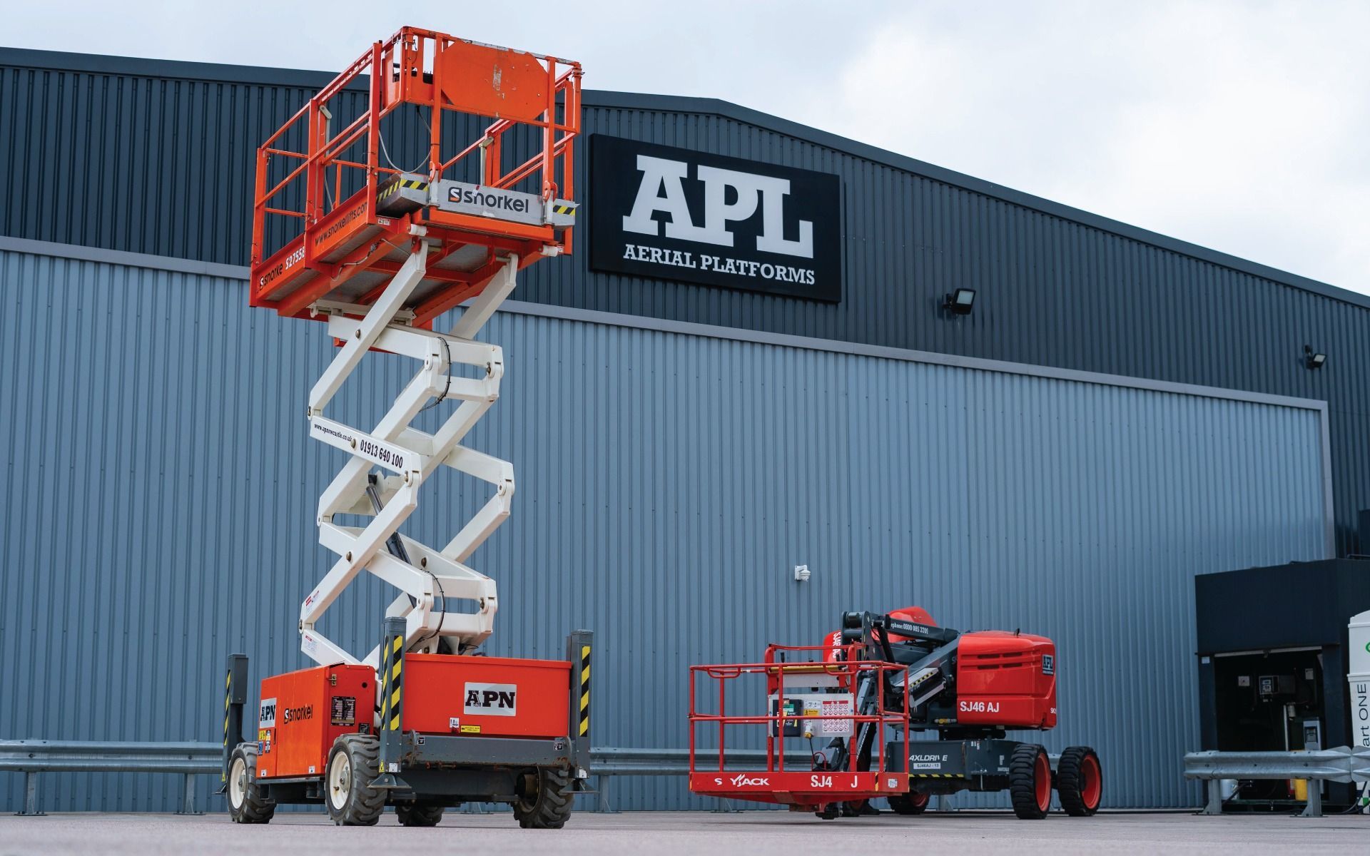 Orange scissor lift extended; red platform lift in front of APL building.