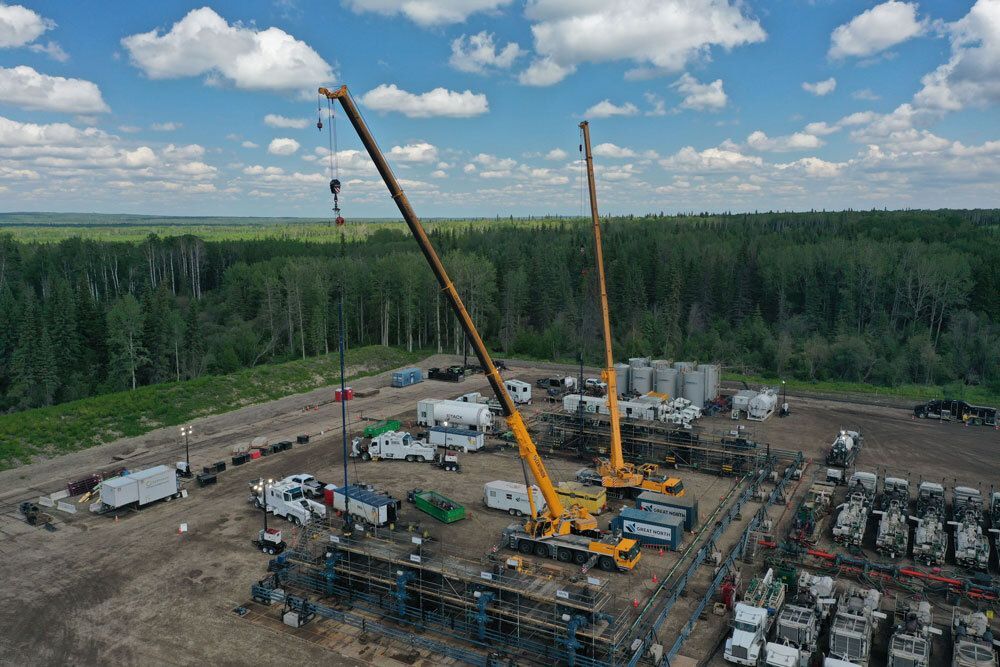 Oil well site with two yellow cranes, trucks, and equipment, surrounded by forest under a blue sky.