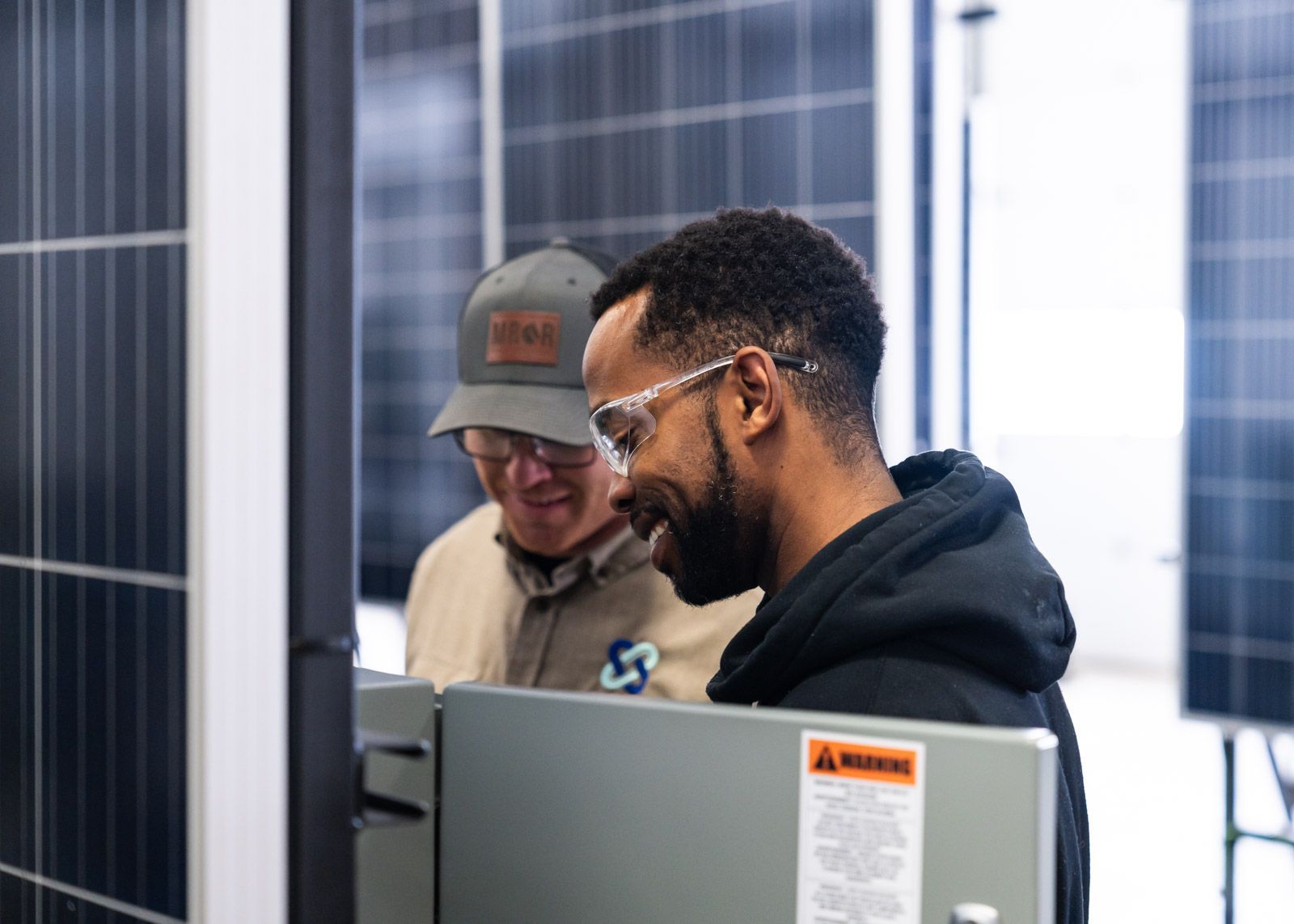 Two men examining electrical equipment near solar panels; one wears a cap and the other safety glasses.