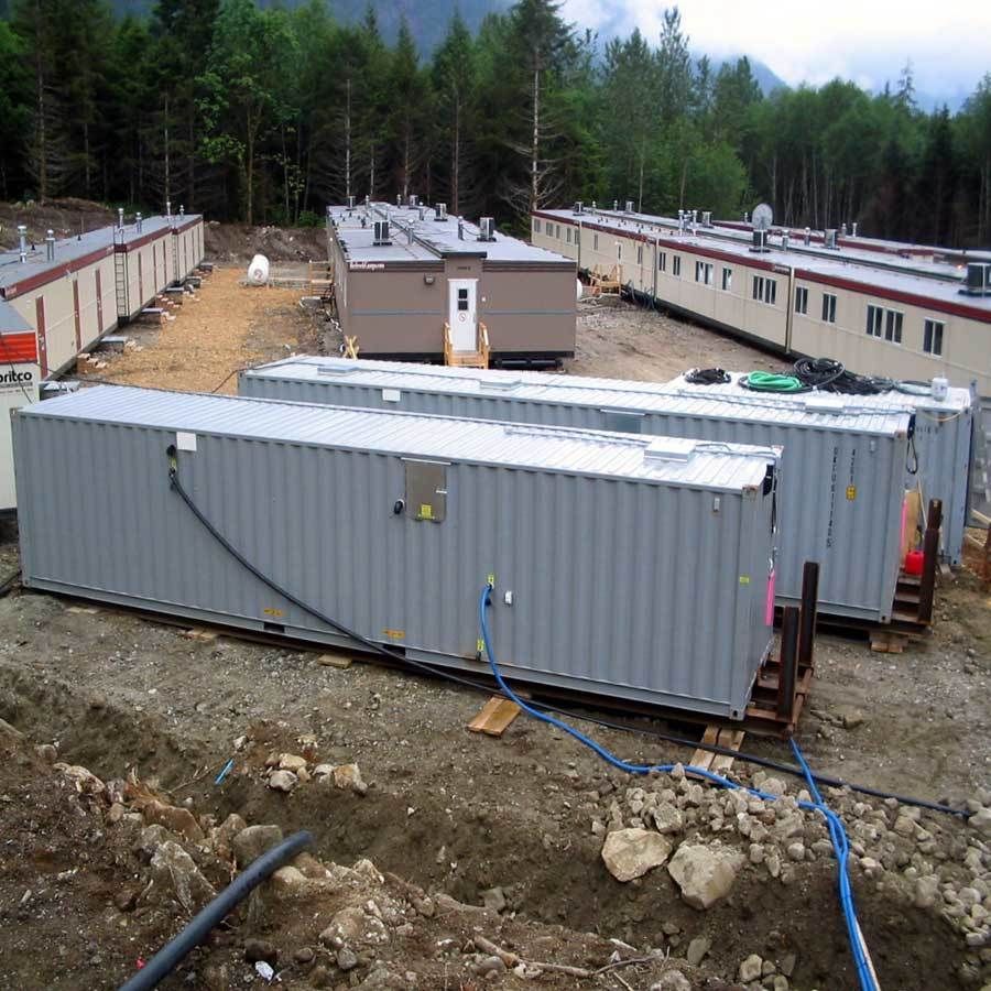 Shipping containers and modular buildings in a wooded area. Gray, brown, and white structures. Blue hoses are visible.