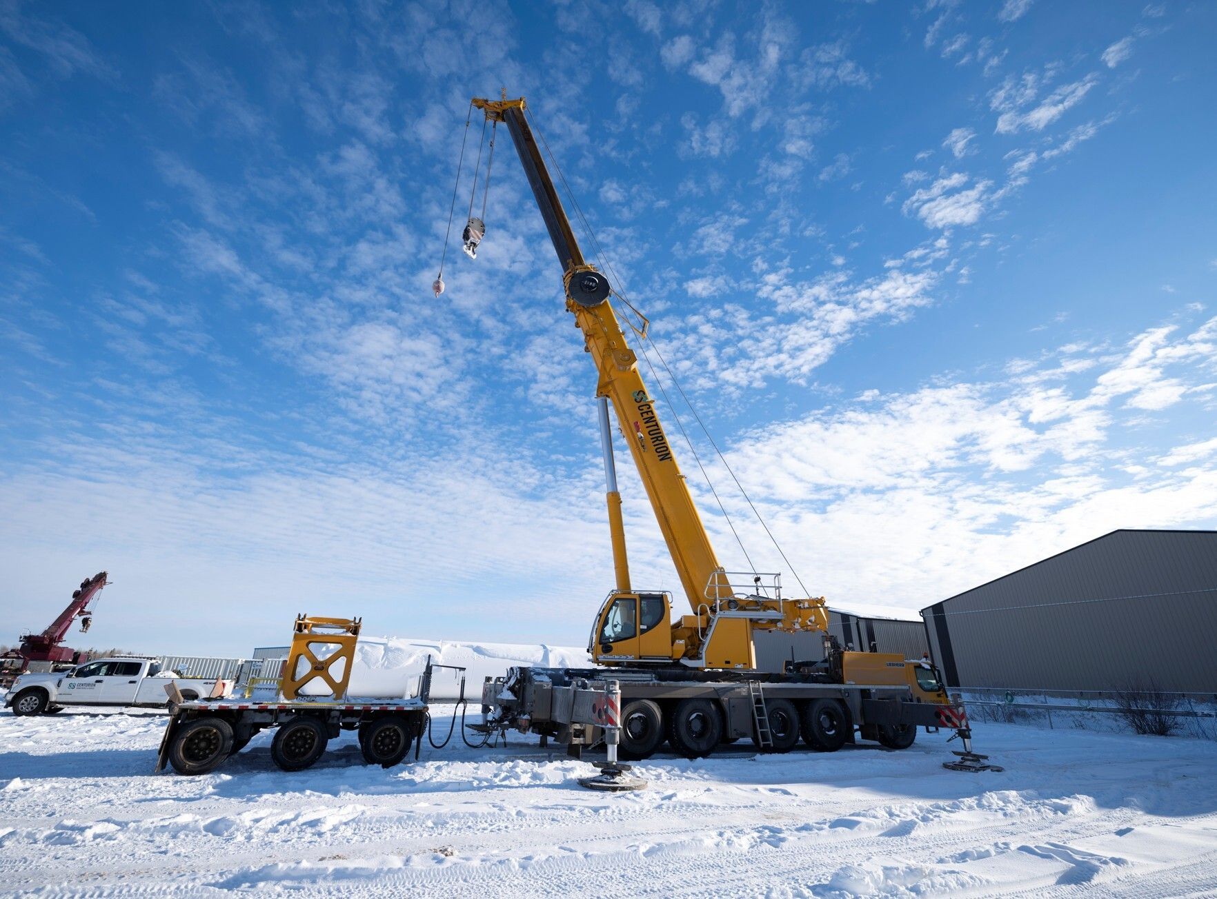Yellow crane on a snowy field, raising equipment. Blue sky with clouds in the background.